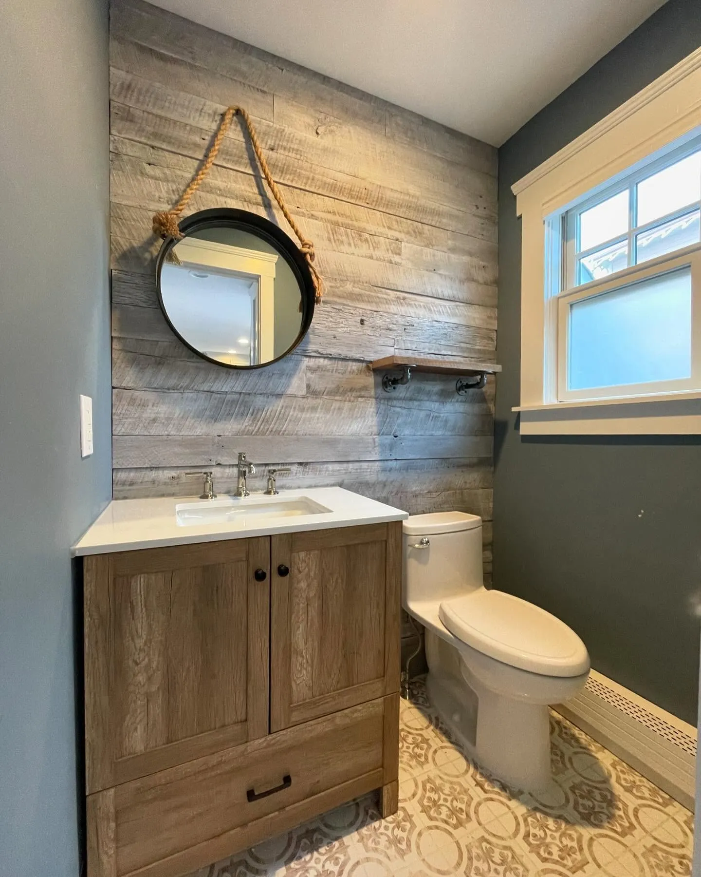 After: the wood-finish vanity, white sink, rustic plank accent wall, round hanging mirror and patterned tile floor.