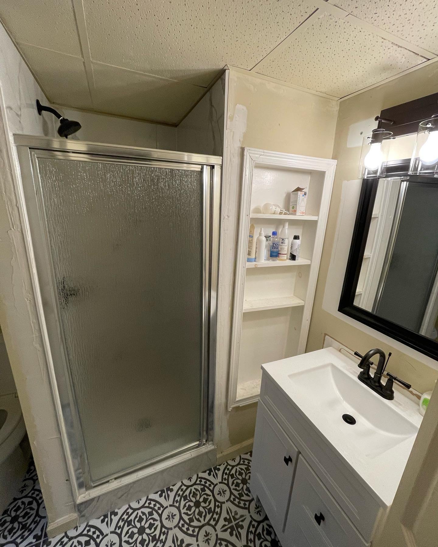 The installed shower, patterned floor, white vanity and vanity light, with wall touch-up areas still visible.