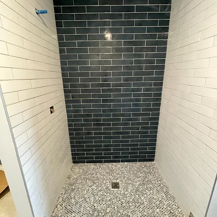 Completed shower tilework: a dark blue back wall, white sidewalls and a light mosaic floor around a square drain.