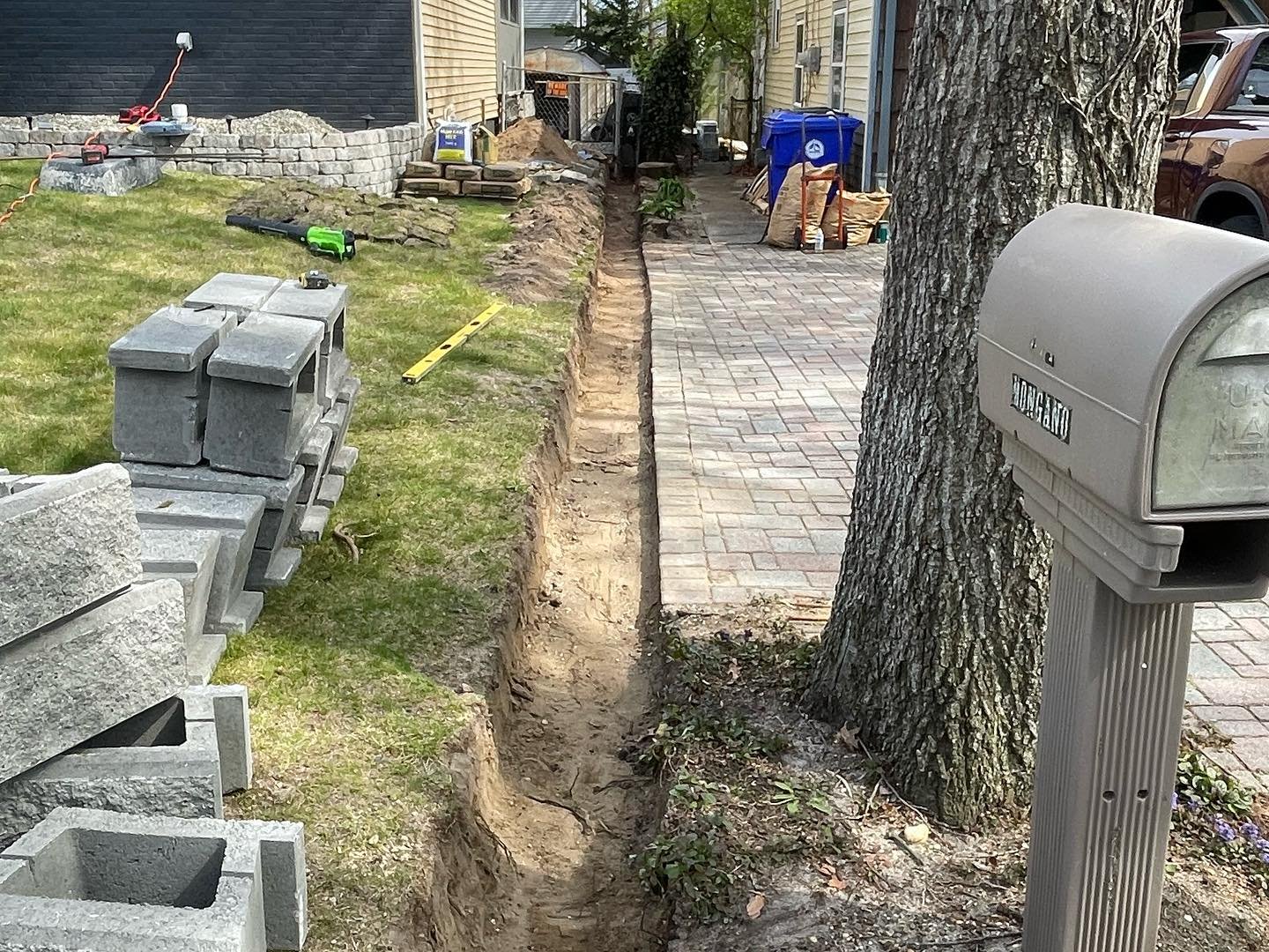 The open wall trench along the lawn, with new blocks delivered and the existing tree and mailbox nearby.